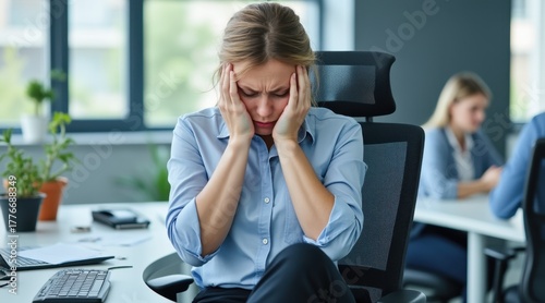 A woman at work appears stressed and overwhelmed, holding her head in frustration amidst a busy office environment.