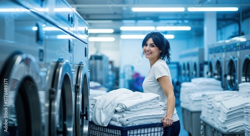 Smiling woman working in an industrial laundry facility. Professional employee pushing a cart with clean white towels in a commercial laundromat