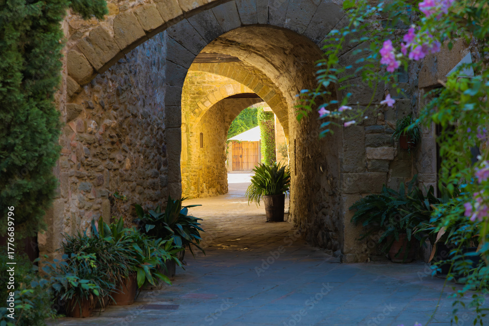 Obraz premium Series of medieval stone arches in Monells, Catalonia, with potted plants and pink flowers creating a romantic and historic atmosphere.