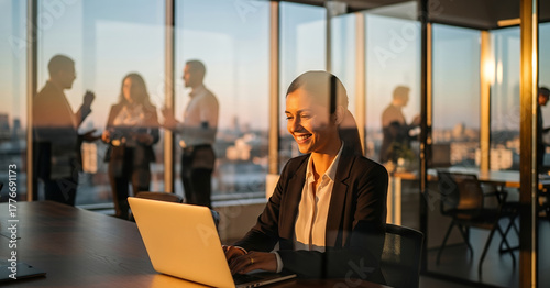 Corporate environment with employees discussing strategy and one focused worker using a laptop by the window at sunset.