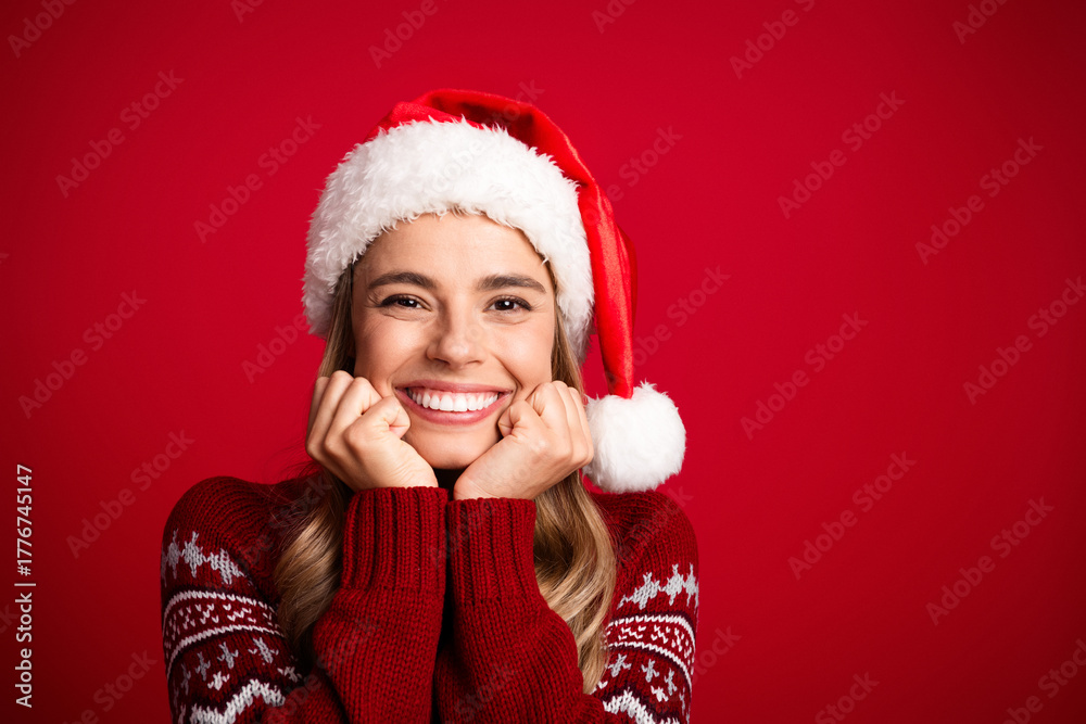 Fototapeta premium Cheerful young woman in a red Christmas sweater and Santa hat smiles brightly against a bold red background perfect for festive holiday stock photos and cheerful seasonal campaigns