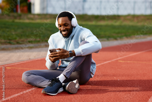 Happy male athlete sits on a running track, smiling while listening to music on his headphones and checking his mobile phone during a workout break.