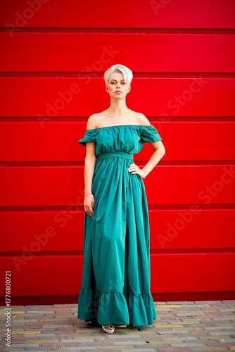 Elegant young woman in a teal off-shoulder maxi dress standing against a vivid red wall. Full-length fashion portrait with strong color contrast and clean, modern background.