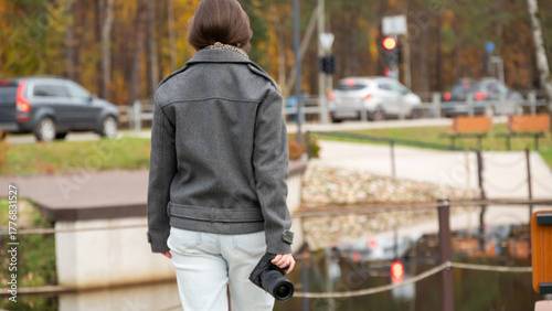 Young Woman Walks Beside Water Holding a Camera in a Park During Autumn Season