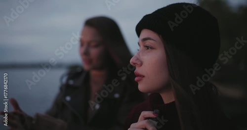 A woman smokes an electronic cigarette near a lake standing with her friend