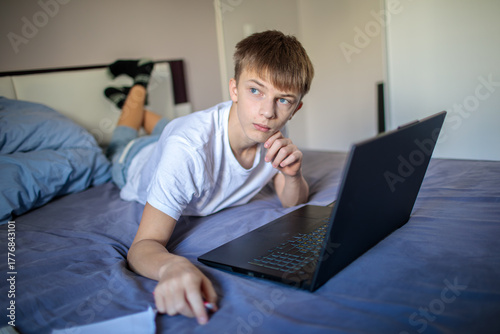 Focused teenage boy studying or working remotely on his laptop while sitting or lying comfortably in bed at home