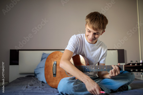 Creative teenage boy sitting on his bed playing an acoustic guitar and writing down lyrics or notes on a notepad