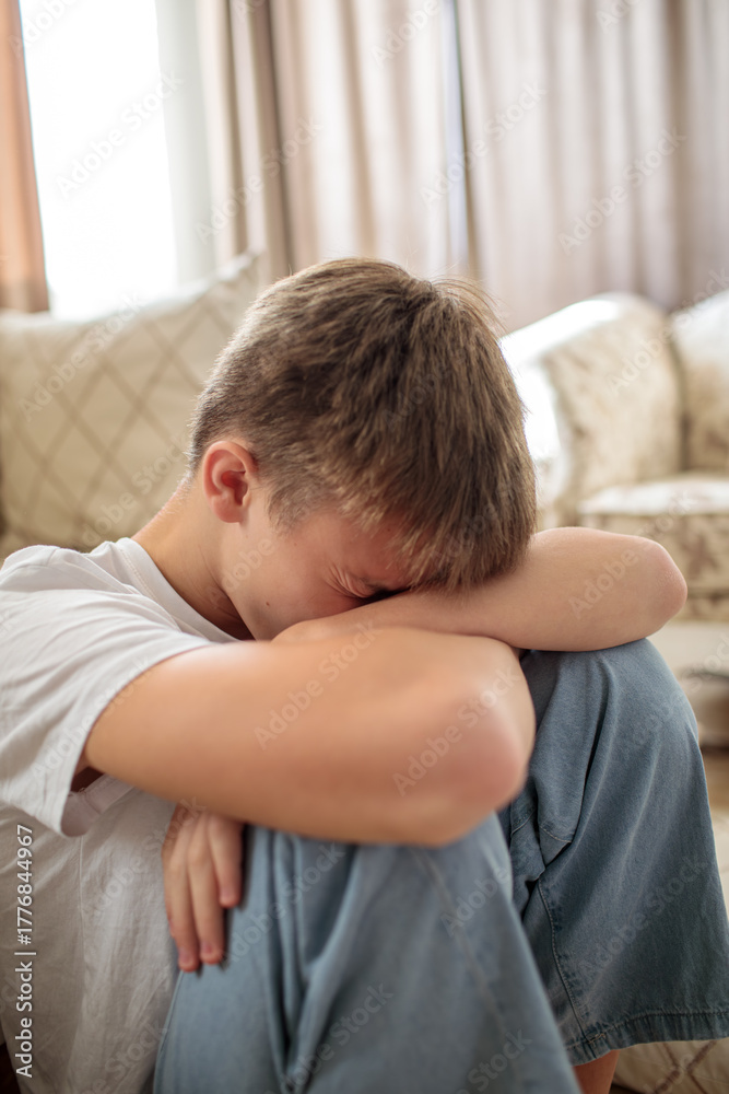 Fototapeta premium Sad and depressed teenage boy sitting on the floor with his head buried in his arms, expressing frustration, anxiety, or mental stress