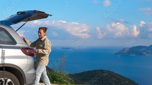 A traveler in a car unloads the trunk. A female traveler unloads things from a car against the backdrop of an endless seascape