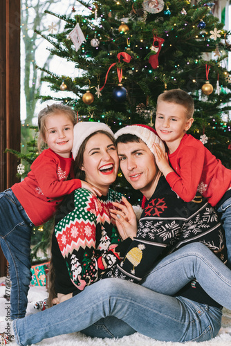 Smiling family portrait in festive sweaters sitting with gifts under Christmas tree at home during holiday. Happy parents and kids cuddling, feeling excited. Family time together. Happy childhood