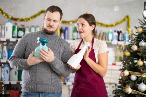 Friendly female store employee in burgundy apron assisting young bearded man selecting cleaning spray in household goods shop adorned with festive ornaments during Christmas season