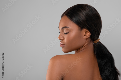 A woman stands with her back turned, her eyes gently closed and a calm expression on her face. Her hair is styled neatly in a ponytail, showcasing natural beauty.