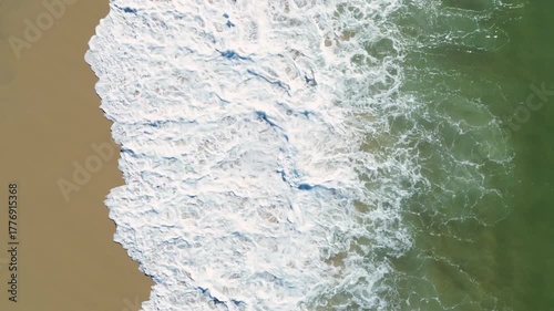 Aerial drone video of waves from the Atlantic Ocean washing onto a sandy beach. Beautiful top view of turquoise water, white sea foam, and golden sand