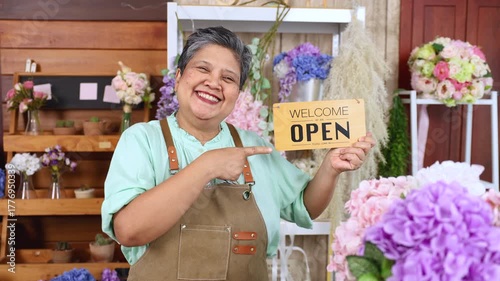 Asian senior woman, florist, small business owner proudly holds open sign with smile, ready to welcome customers to flower shop, concept of new beginning, retail startup, elderly with passion success