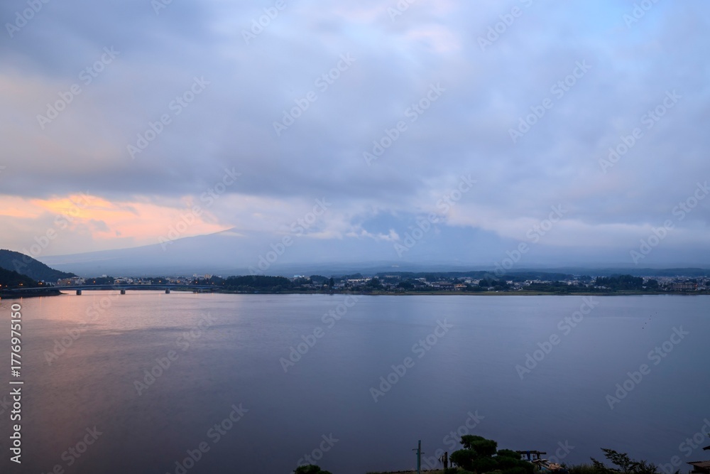 Fototapeta premium Lakeside Cityscape at Sunrise with Bridge and Mountain Backdrop Beneath Dramatic Cloud Cover