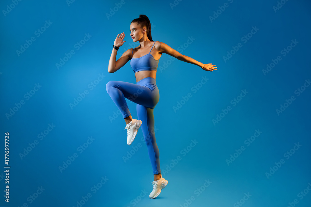 Naklejka premium A female athlete is executing a powerful jump while training in a blue studio background. She appears focused and energized, embodying the spirit of fitness and motivation.