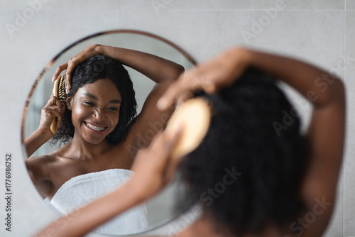 Photos A cheerful black lady with bushy hair brushes her hair in front of a mirror