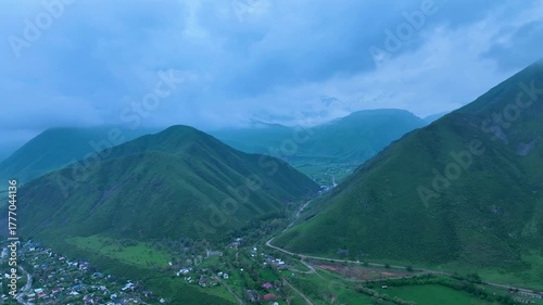 Aerial view of a small village winding along the bottom of a deep, green, steep-sided mountain valley under a dramatic, cloudy blue sky at twilight