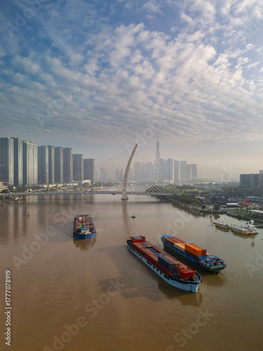 Transporting containers on barges Saigon River early sunny clear morning with Ho Chi Minh City suspension bridge, modern architecture and landmark in the background