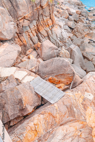 Wooden Walkway Amidst the Rocks: The image captures a wooden walkway delicately placed amidst the rugged embrace of towering rocks. Coastal rocks in soft pastel tones