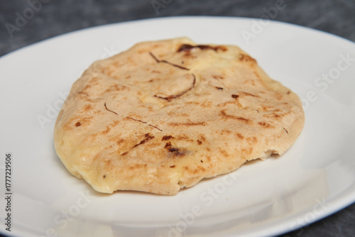 A close-up photo of a golden, cheese-filled pupusa on a white plate against a black-grey marble background.