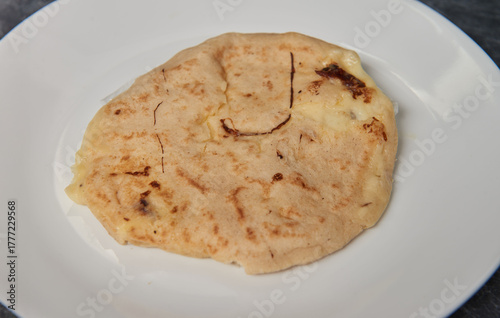 A close-up photo of a golden, cheese-filled pupusa on a white plate against a black-grey marble background.