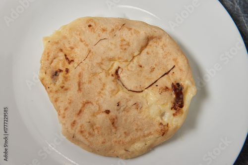 A close-up photo of a golden, cheese-filled pupusa on a white plate against a black-grey marble background.