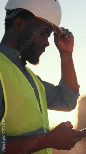 African american builder using smartphone in construction site, vertical portrait outdoors. Black man with hard hat for protective head, internet addiction, new mobile app and software for work