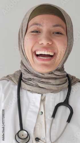 Vertical video. Cheerful young muslim doctor in hijab and lab coat laughing confidently with stethoscope, professional healthcare portrait on white background