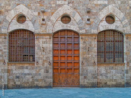 Symmetrical brick façade in Siena, Italy, with a central wooden door flanked by two arched windows with grilles. The warm tones of stone and craftsmanship evoke the elegance of Tuscan architecture.