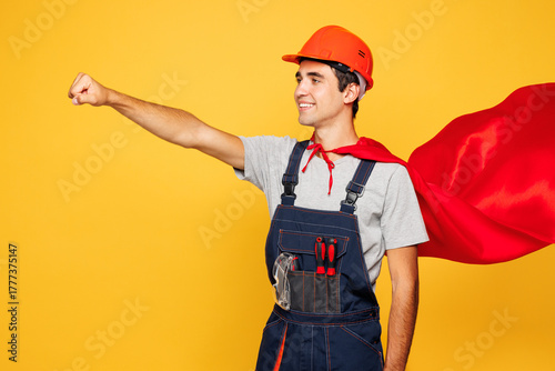 Obraz na plátně Young employee laborer handyman man wear helmet overall red coat do super hero gesture isolated on plain yellow background
