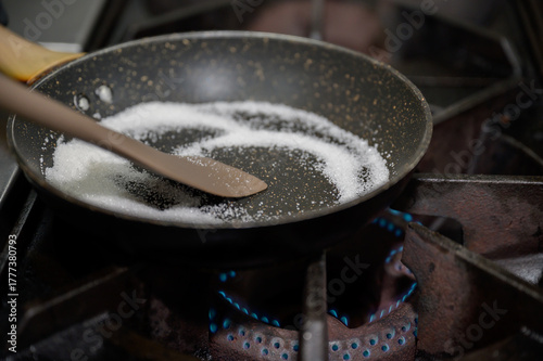Close-up shot of sugar melting in nonstick pan over blue gas flame, concept of dessert preparation and caramel making in professional kitchen.