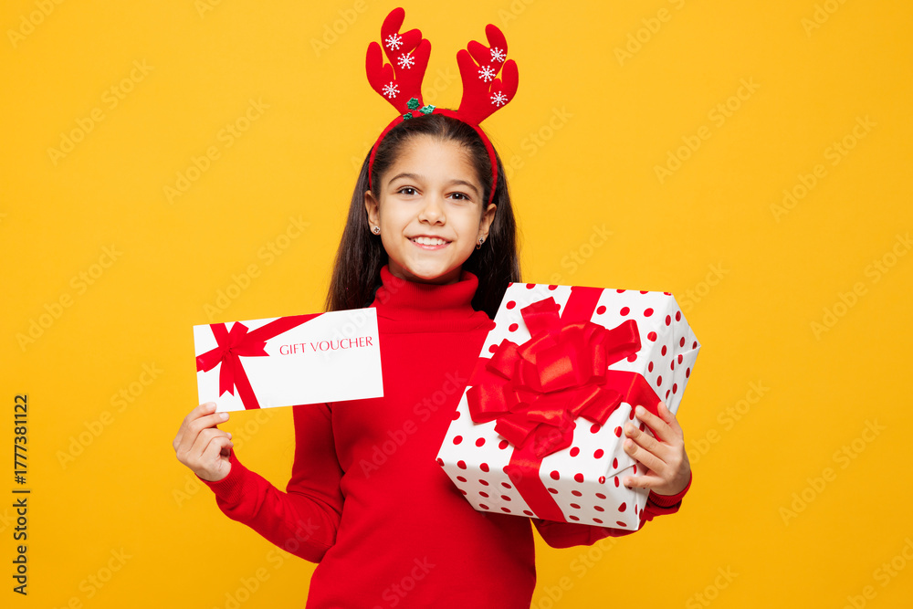 Fototapeta premium Merry little girl wear red clothes decorative deer horns on head posing hold present box store gift coupon voucher card isolated on plain yellow background. Happy New Year Christmas holiday concept.