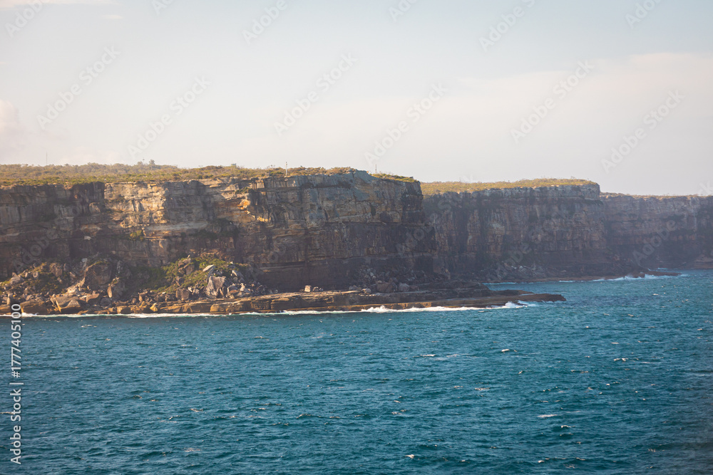 Fototapeta premium High-quality photo of Sydney’s coastal cliffs near Manly, captured from a unique vantage point on a cruise ship at golden hour. The image showcases rugged rock formations and bright blue ocean water