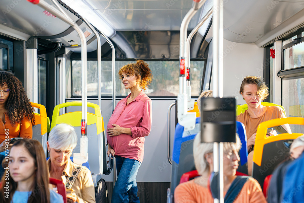 Obraz premium Pregnant woman commuting on a city bus, gently touching her belly while standing among other passengers during daily travel