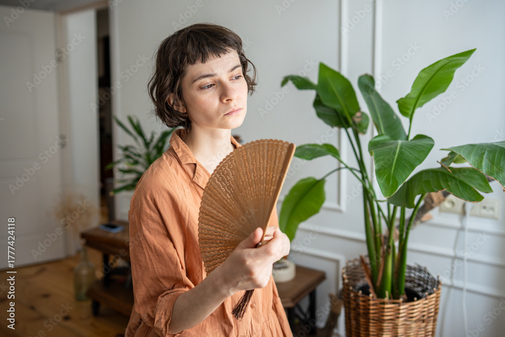 Obraz premium Woman at home suffering from summer heat, standing beside window in living room, waving hand paper fan for cooling down. Tired young female with sick expression, overwhelmed by summer heat or fever.
