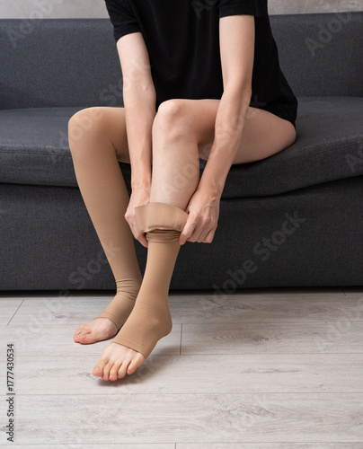 Faceless female wearing compression socks sitting on grey sofa indoors demonstrating healthy lifestyle, vein care and prevention of venous disease