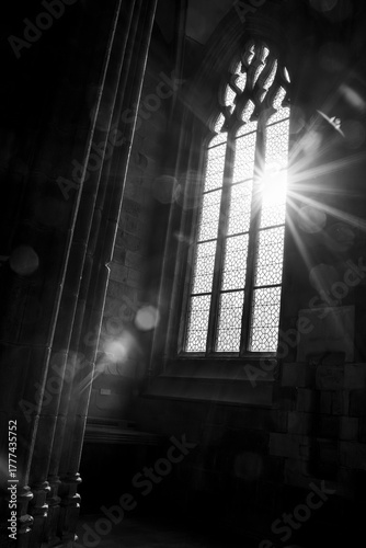 Black and white image of gothic arched stained glass windows inside Mont Saint Michel, with sunlight beaming through the glass and casting rays across the historic stone architecture

