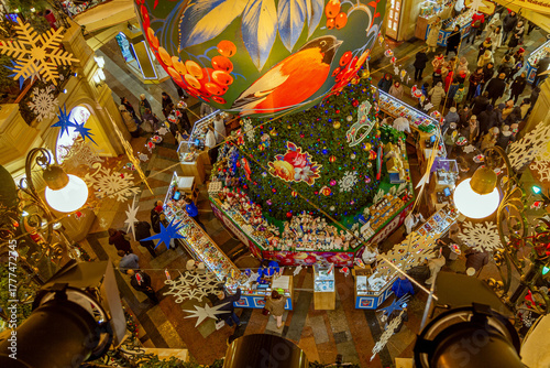 An aerial view of a festive Christmas market in a decorated shopping mall, complete with a large tree, ornaments, snowflakes, and shoppers.