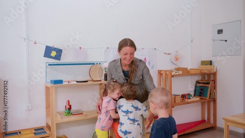 Smiling teacher embraces small children in cheerful classroom surrounded by shelves, drawings, and learning materials, expressing affection, care, and connection during joyful preschool