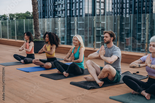 Multinational group of people practices yoga on mats with city skyline and palm tree under sky. Hands in prayer suggest guided meditation during yoga class
