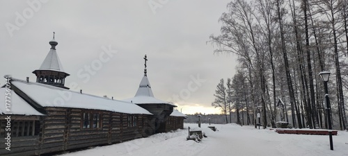 A wooden fence. A winter day, the wooden monastery buildings are covered in snow. The rough-hewn wood has been blackened by time. Tall trees grow opposite. White snow lies all around.