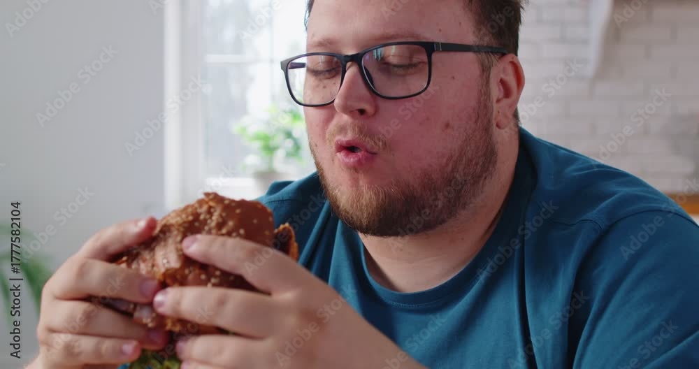 Obese chubby man holding burger to bite with appetite, eating fat fast food at home kitchen. Unhealthy diet choice for lunch and dinner of overweight person with obesity, big belly and glasses