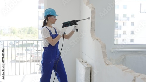 Woman construction worker demolishing interior wall with rotary hammer, wearing safety overalls, glasses and hardhat, is creating substantial concrete debris pile during demolition process
