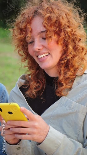 A cheerful girl with curly hair is happily using her smartphone to connect with friends on social media, showcasing modern communication and the joy of sharing experiences through technology.