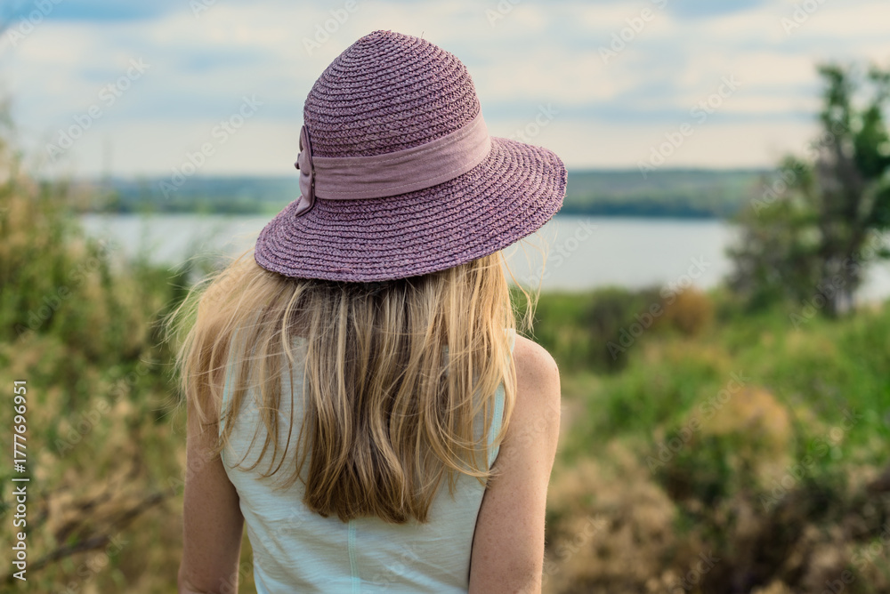 Φωτογραφία Back view of blonde woman in summer hat looking at scenic view of lake
