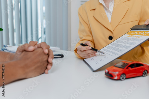 A car sales negotiation takes place in an office where a female salesperson in a yellow blazer explains details of a contract to a male client, showing car models and handing over keys.