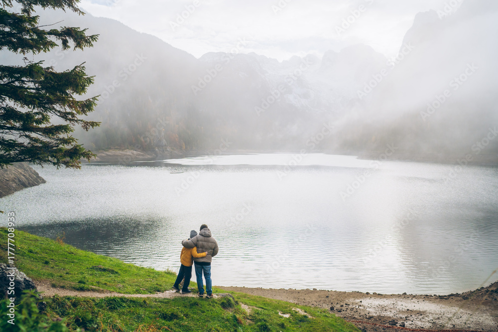 Obraz premium Father and son enjoy a peaceful moment by the serene lake surrounded by mountains on a foggy day