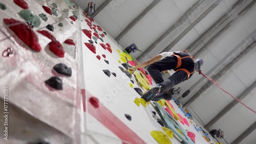 Two people practicing indoor rock climbing routes at an artificial gym wall, one person wearing a harness with rope and another person without a harness engaged in bouldering training