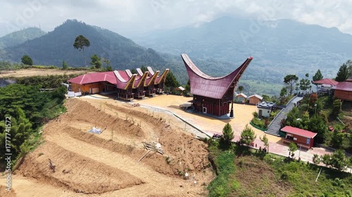 Aerial view of traditional Tongkonan houses with curved roofs on a hillside in Tana Toraja, Indonesia, surrounded by mountains and clouds.	
